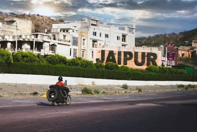 Man on bike carrying tires passing Jaipur city sign under cloudy sky, Jaipur tour package